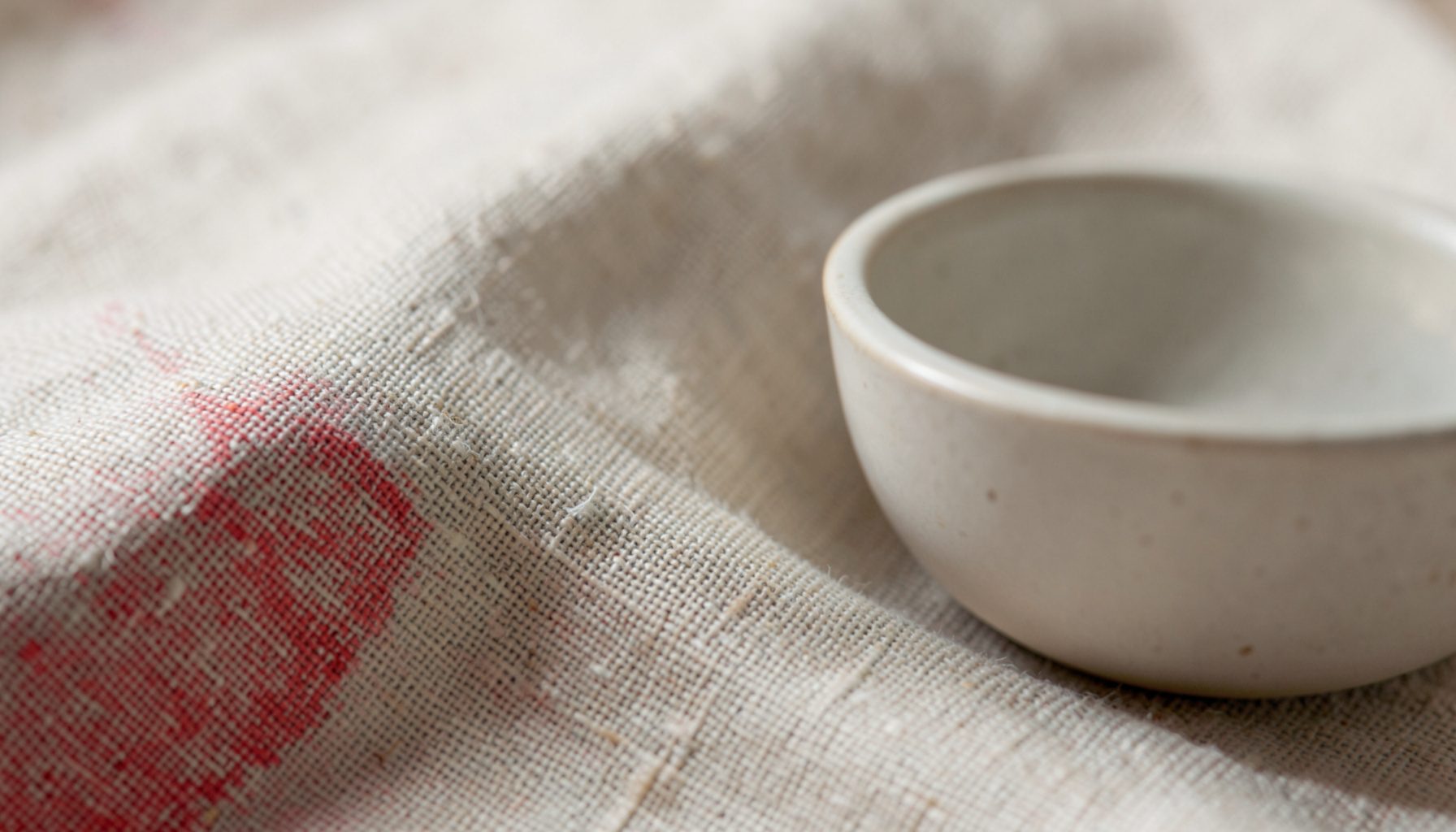 An extreme close-up photograph of textured natural linen fabric beside a simple ceramic bowl, soft cream and beige tones with gentle shadows