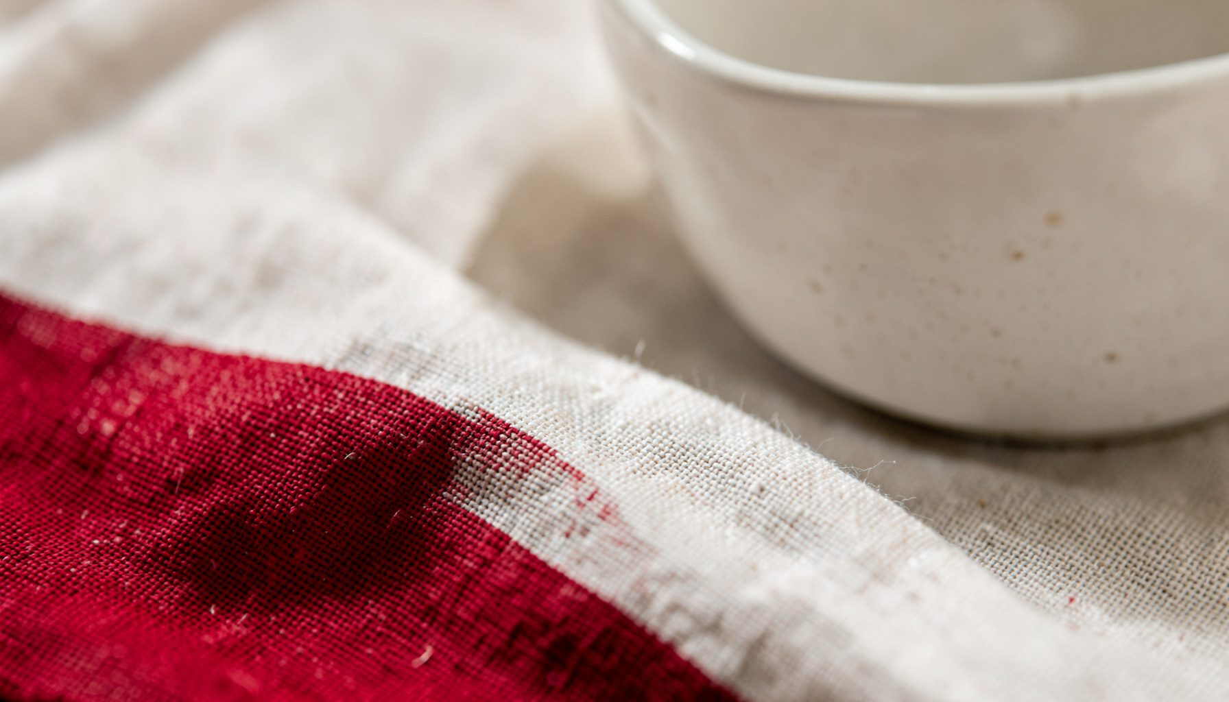 Extreme close-up of a textured linen napkin beside a ceramic bowl, soft cream and beige tones with gentle shadows